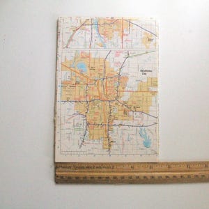 May include: A vintage map of Oklahoma City, showing a detailed road network in red, orange, and blue. The rectangular map has the words "Oklahoma City" printed on it. A wooden ruler is placed at the bottom, showing inch measurements.