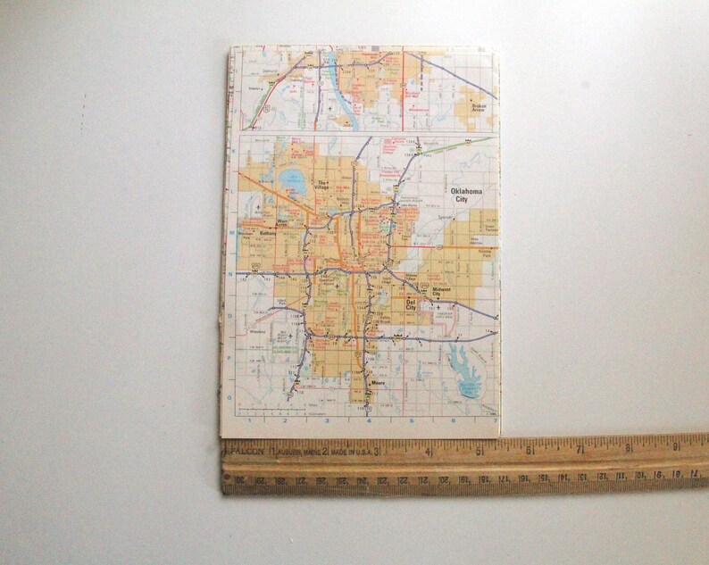 May include: A vintage map of Oklahoma City, showing a detailed road network in red, orange, and blue. The rectangular map has the words "Oklahoma City" printed on it. A wooden ruler is placed at the bottom, showing inch measurements.