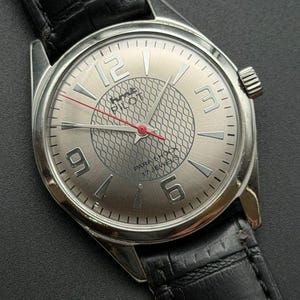 Puede incluir: Reloj de pulsera plateado con una correa de cuero negro. La esfera del reloj presenta grandes números plateados en las posiciones de las 12, 3, 6 y 9 en punto. El reloj tiene un centro texturizado y la palabra "Pilot" es visible.