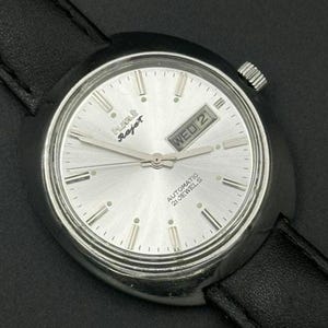 May include: Silver-faced wristwatch with a black leather strap. The watch shows the day and date, with "WED 2" visible. The watch also features the text "AUTOMATIC 21 JEWELS" and the brand name "HMT Rajat".