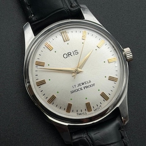 May include: A vintage Oris wristwatch with a silver sunburst dial, gold-tone hour markers, and gold-tone hands. The dial features the text ORIS, 17 JEWELS, SHOCK PROOF, and SWISS MOVT. It has a black leather strap with a textured crocodile-style pattern.