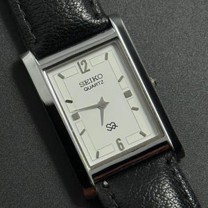 May include: A rectangular Seiko Quartz watch with a white face, black hands, and silver-tone case. The watch has a black leather strap. The watch face displays the number 12 and 6, with the Seiko Quartz logo.