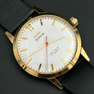 May include: A gold wristwatch with a white face, gold hands, and a red second hand. The watch face displays the text "SONA" and "PARA SHOCK 17 JEWELS". It is set on a black leather strap.