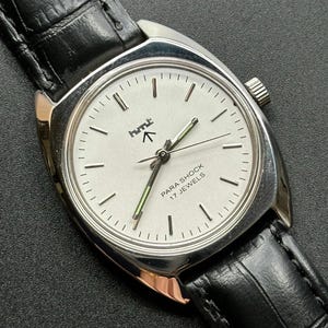 May include: A vintage wristwatch with a silver-tone case and a white face. The watch features black hour markers, a second hand with a green tip, and the text "PARA SHOCK 17 JEWELS". It has a black leather band.