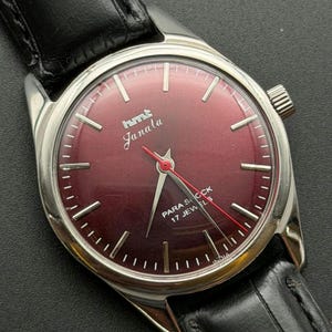 Peut inclure: Une montre-bracelet vintage avec un cadran bordeaux et un boîtier argenté. La montre affiche le texte "hmt Janala" et "Para Shock 17 Jewels". Elle est dotée d'un bracelet en cuir noir et d'une couronne argentée. Les aiguilles sont argentées et rouges.