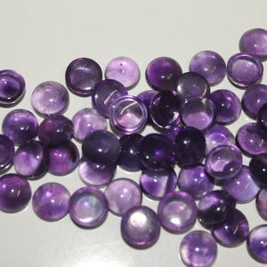 Può includere: Una collezione di cabochon rotondi in ametista lucida in varie tonalità di viola. Le pietre preziose sono sparse su una superficie chiara, mostrando le loro qualità traslucide e opache. Questi cabochon sono ideali per la creazione di gioielli e l'artigianato.