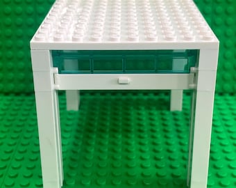 May include: A white plastic building block table featuring a flat top surface with circular studs and a translucent teal rectangular panel integrated into the front frame, supported by four vertical white legs constructed from interlocking bricks.