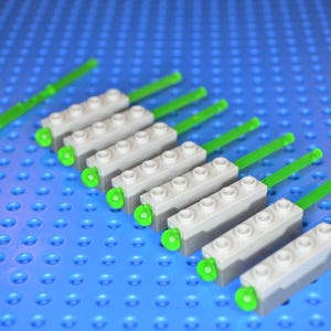 May include: A collection of seven grey rectangular building blocks, each featuring four studs on top and a long, translucent neon green antenna piece extending from one side, arranged in a diagonal row on a blue studded base plate.
