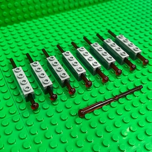 May include: An overhead shot shows several gray Lego bricks with dark red rods and red translucent tips, arranged on a green Lego base. The Lego bricks are rectangular with circular studs. One rod is separate from the others.