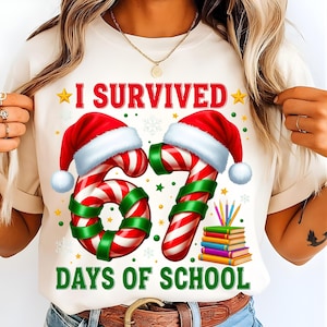 Puede incluir: Camiseta color crema con el texto "I Survived 67 Days of School". El diseño presenta bastones de caramelo con gorros de Papá Noel, detalles verdes, estrellas, copos de nieve y una pila de libros con lápices. Un diseño festivo y de celebración.