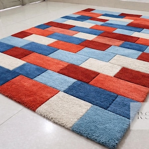 Può includere: Tappeto rettangolare con motivo geometrico. Il tappeto presenta un design patchwork di forme rettangolari nei toni del rosso, blu e bianco sporco. Le dimensioni del tappeto non sono visibili nell'immagine.