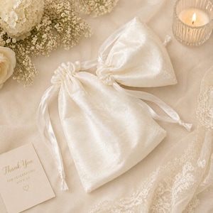 May include: Ivory satin drawstring bags with matching ribbon ties, arranged on a cream-colored surface. A small card with the words "Thank You" is visible. The scene is decorated with white flowers and a lit candle.