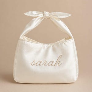 Puede incluir: Un bolso de mano de satén color crema con un asa superior anudada y el nombre sarah bordado en un color de hilo a juego en la parte delantera. El bolso tiene una base rectangular estructurada con esquinas redondeadas y un acabado brillante.