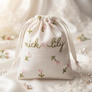 May include: A cream-colored drawstring pouch featuring embroidered pink flower buds with green stems and leaves. The text "nick + lily" is stitched in green thread across the center, with a small pink heart embroidered between the two names.