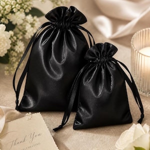 May include: Two black satin drawstring bags of different sizes. The bags have a shiny finish and are gathered at the top with matching black ribbons. A small card with the words "Thank You" is visible in the foreground.