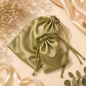 May include: Two olive green satin drawstring bags of different sizes. The bags have a shiny, smooth texture and are gathered at the top with matching drawstrings. The background includes a cream-colored satin fabric and sprigs of dried flowers.