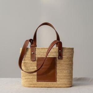 Könnte beinhalten: Eine rechteckige Stroh-Tragetasche mit braunen Lederdetails. Die Tasche hat einen braunen Lederaufnäher auf der Vorderseite und braune Ledergriffe. Die Tasche besteht aus gewebtem Stroh und hat ein schlichtes, elegantes Design.
