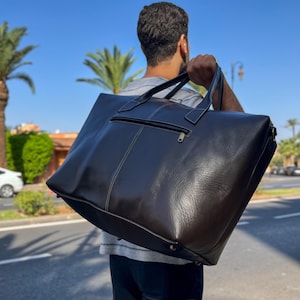 May include: A large, black leather tote bag with a zip and white stitching. The bag has two handles and a shoulder strap. The bag is being carried outdoors on a sunny day.