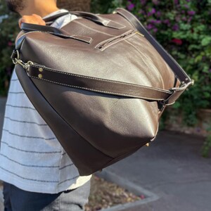 May include: A large, dark brown leather tote bag with a shoulder strap and white stitching. The bag has a zippered pocket and metal hardware. The bag is carried over the shoulder.