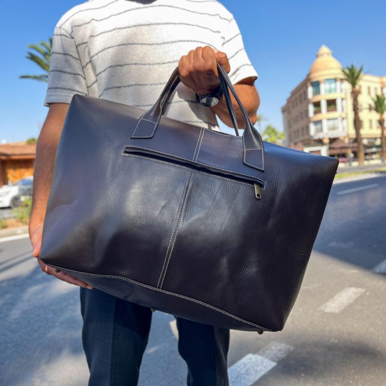 May include: A large, dark brown leather tote bag with white stitching and a zipped pocket. The bag has two handles. The rectangular bag is held by a person. The bag appears to be new.