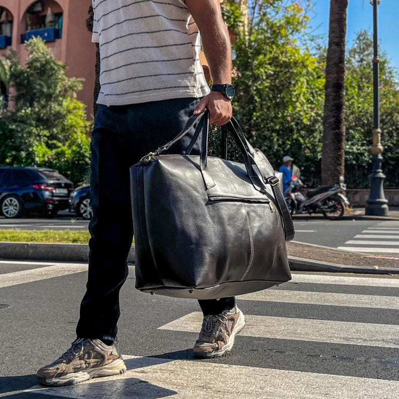 May include: A large, dark brown leather travel bag with a top handle and shoulder strap. The bag has a rectangular shape and a zippered pocket on the front. The bag is being carried across a pedestrian crossing.