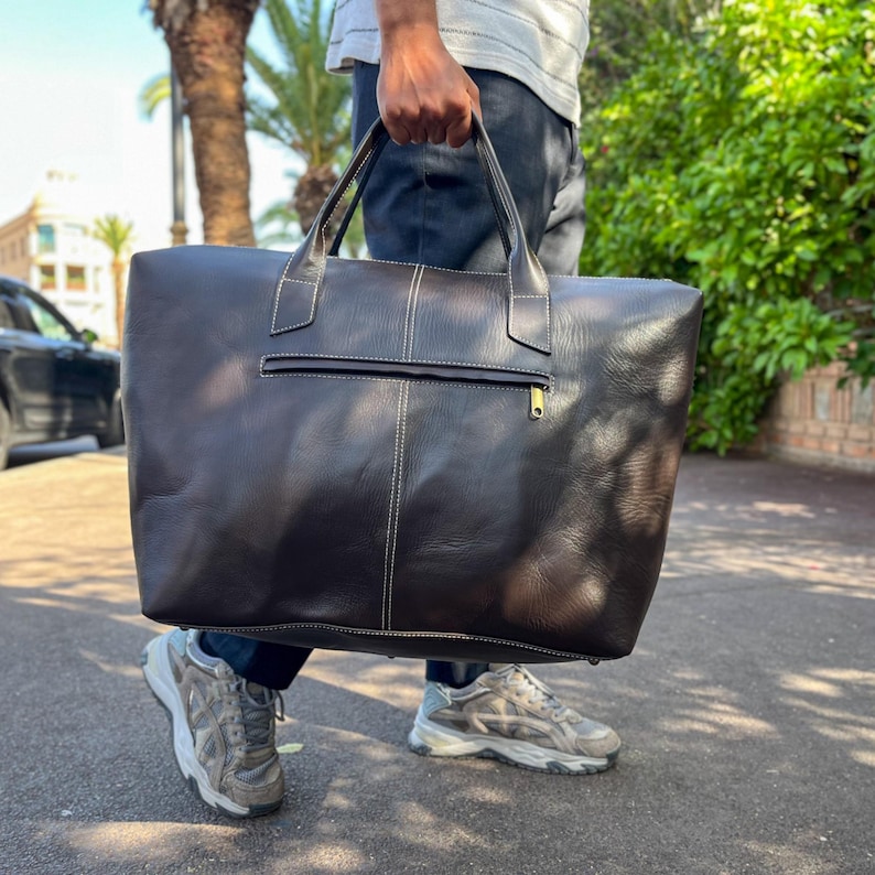 May include: A dark brown leather travel bag with white stitching and a zipped pocket. The bag has two handles and a gold-coloured zip. The bag is being held by a person.