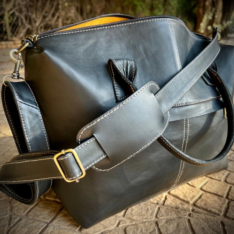 May include: Black leather duffel bag with white stitching and a yellow interior. The bag features a shoulder strap with a padded section and a gold-coloured buckle. The bag has a zip closure and a handle on top.