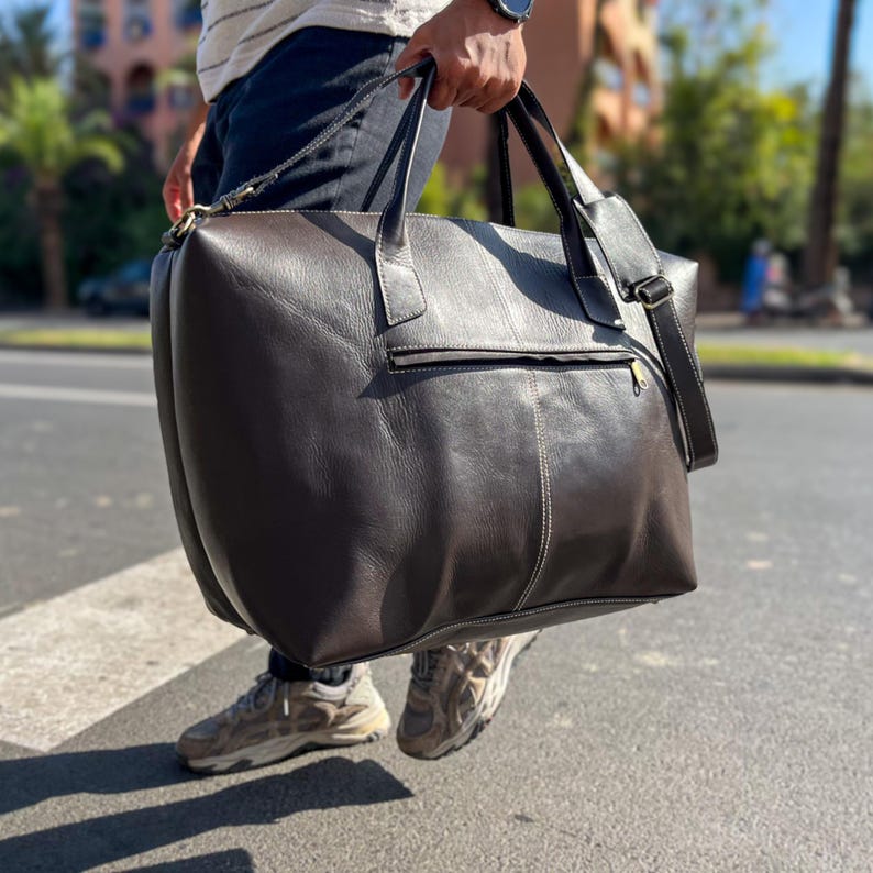 May include: A dark brown leather travel bag with a top handle and a shoulder strap. The bag has a rectangular shape with a zippered front pocket and visible stitching. The bag is being carried outdoors.