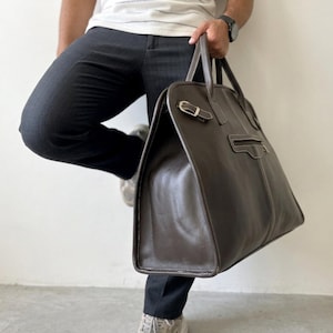 May include: A brown leather tote bag with a rectangular shape and a top handle. The bag features a buckle closure and white stitching details. The bag is held by a person wearing a white shirt and black pants.