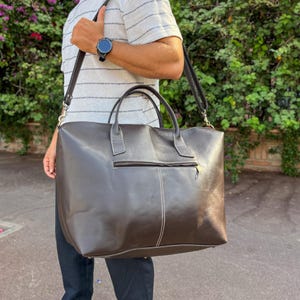 May include: A large, dark brown leather tote bag with a shoulder strap. The bag has a rectangular shape with a zippered front pocket and white stitching details. The bag is carried over the shoulder.