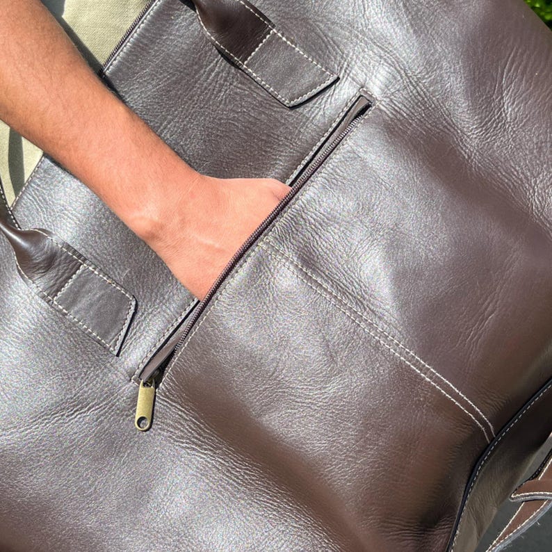 May include: A brown leather tote bag with a zipped pocket. The bag features white stitching and a brass zip. The handle is visible on the left. A hand is reaching into the zipped pocket.