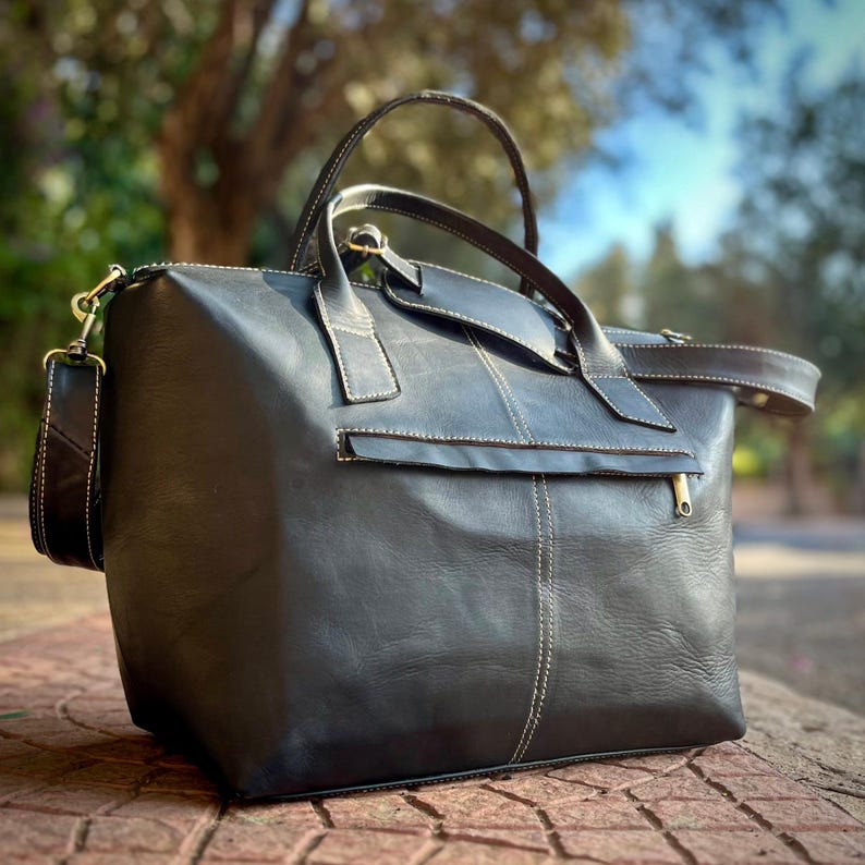 May include: A black leather travel bag with a top handle and a detachable shoulder strap. The bag has white stitching, a front zipped pocket, and a gold-coloured zip. The bag is on a textured surface.