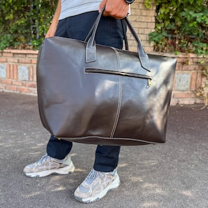 May include: A large, dark brown leather duffel bag with white stitching and a zippered pocket. The bag has two handles and a shoulder strap. The bag is being held by a person wearing dark blue jeans and grey trainers.