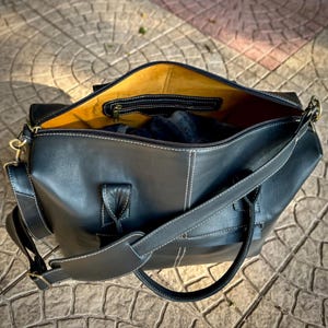 May include: Black leather duffel bag with white stitching and a yellow interior. The bag has a zippered pocket inside and a detachable shoulder strap. The bag is open, revealing a glimpse of the contents.