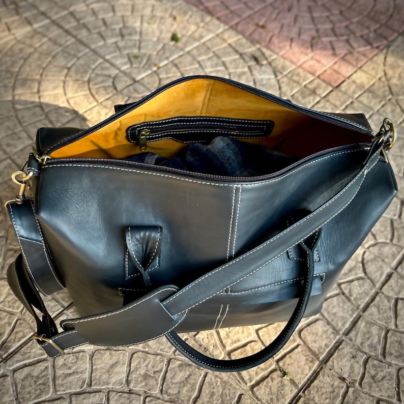 May include: Black leather duffel bag with white stitching and a yellow interior. The bag has a zippered pocket inside and a detachable shoulder strap. The bag is open, revealing a glimpse of the contents.