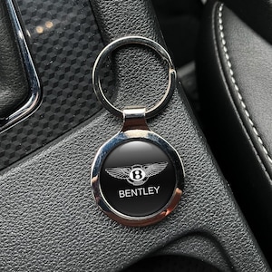 Könnte beinhalten: Ein silberner Bentley-Schlüsselanhänger mit einem schwarzen kreisförmigen Emblem mit dem Bentley-Logo und dem Wort "BENTLEY" in Weiß. Der Schlüsselanhänger ist rund und an einem silbernen Schlüsselring befestigt.