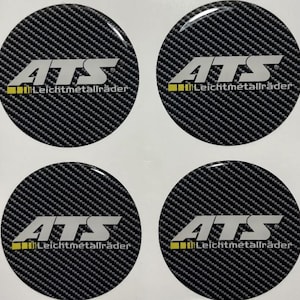 Puede incluir: Cuatro tapacubos circulares para ruedas de coche con un patrón de fibra de carbono. Cada tapón presenta las letras blancas "ATS" sobre el texto "Leichtmetallräder" en blanco y amarillo.