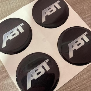 Puede incluir: Cuatro pegatinas circulares negras con el logotipo ABT, acabado brillante. El logotipo ABT es plateado, con una fuente moderna y en negrita. Las pegatinas están sobre un soporte blanco.