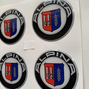Puede incluir: Cuatro pegatinas circulares con el logotipo de Alpina. Cada pegatina presenta un diseño de escudo dividido en secciones rojas y azules, con un borde plateado y negro. La palabra "ALPINA" está impresa en plata alrededor del borde de cada pegatina.