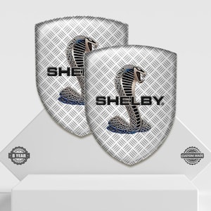 May include: Two shield-shaped emblems with a silver diamond plate pattern. Each emblem features a cobra design in black, blue, and silver, with the word "SHELBY" in black block letters. The emblems are displayed on a white surface.