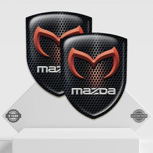 May include: Two black shield-shaped decals with a perforated texture. Each features a red Mazda logo and the brand name in white. The decals are displayed on a white surface.