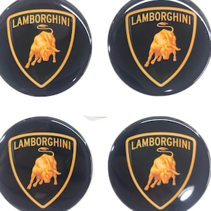 Puede incluir: Cuatro emblemas circulares negros con el logotipo de Lamborghini. Cada emblema tiene un escudo dorado con un toro dorado y la palabra "Lamborghini" en letras doradas. Los emblemas son brillantes y parecen estar hechos de un material duradero.