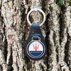 May include: Black leather Maserati keyring with a silver key ring. The keyring features a circular emblem with the Maserati logo: a red trident over a white background, with a blue border and the name "MASERATI".