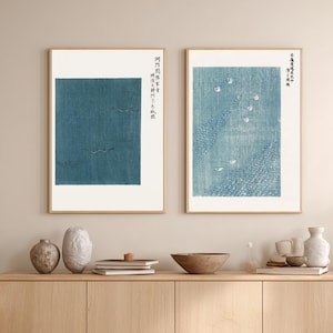 Puede incluir: Dos grabados enmarcados con obras de arte de estilo japonés. Uno presenta un diseño rectangular azul turquesa con pájaros en vuelo. El otro grabado tiene un diseño azul y blanco con un río y pájaros. Ambos grabados tienen marcos dorados y texto japonés.