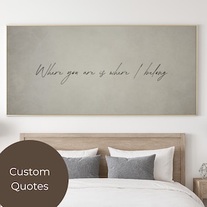 Op de afbeelding: Een ingelijst kunstwerk met de quote "Where you are is where I belong" in elegant schrift. Het kunstwerk heeft een neutrale, getextureerde achtergrond en een gouden lijst. De afbeelding wordt weergegeven boven een bed met kussens.