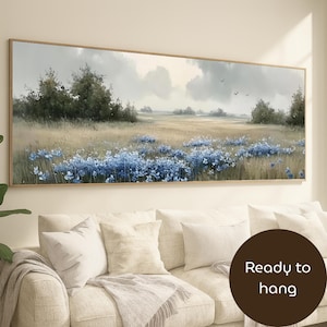 May include: A framed landscape painting depicts a field of blue flowers under a cloudy sky. The artwork features a muted color palette with shades of green, beige, and blue. The painting is ready to hang and is displayed above a white sofa.