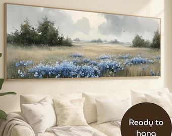 May include: A framed landscape painting depicts a field of blue flowers under a cloudy sky. The artwork features a muted color palette with shades of green, beige, and blue. The painting is ready to hang and is displayed above a white sofa.