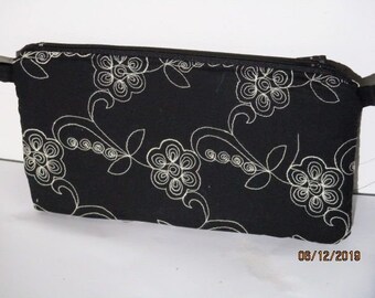 Op de afbeelding: Zwarte ritszak met een wit bloemenontwerp. De tas is gemaakt van een zachte stof en heeft een ritssluiting.