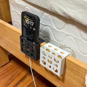 May include: A black phone holder with a phone displaying the time 10:17, and a white holder with circular cutouts, both attached to a wooden bed frame. A white charging cable is connected to the phone.