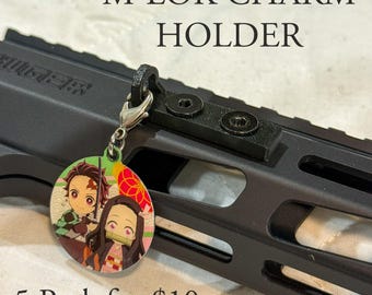 Könnte beinhalten: Schwarzer M-LOK-Charm-Halter mit einem runden Charm mit Anime-Charakteren. Der Charm ist mit einem silbernen Verschluss befestigt. Der Text "M-LOK CHARM HOLDER" befindet sich über dem Produkt.